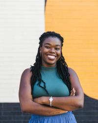 Golden Owens, a black woman with waist-long hair, stands with arms crossed, smiling in front of a black, white, and yellow mural.  Prof Owens wears a teal tank top and blue pants with a watch and hoop earrings.