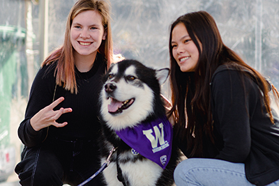 Two students kneel next to dubs the husky