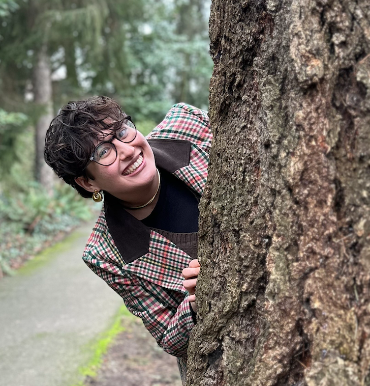 Picture of Rachel, a white passing femme with short brown hair and brown eyes.  Rachel is wearing a brown, pink, and green checked blazer and glasses while popping out from behind a tree.